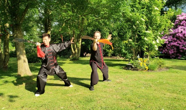 Zwei Personen in traditionellen Kostümen zeigen Kampfkunst in einem Garten.