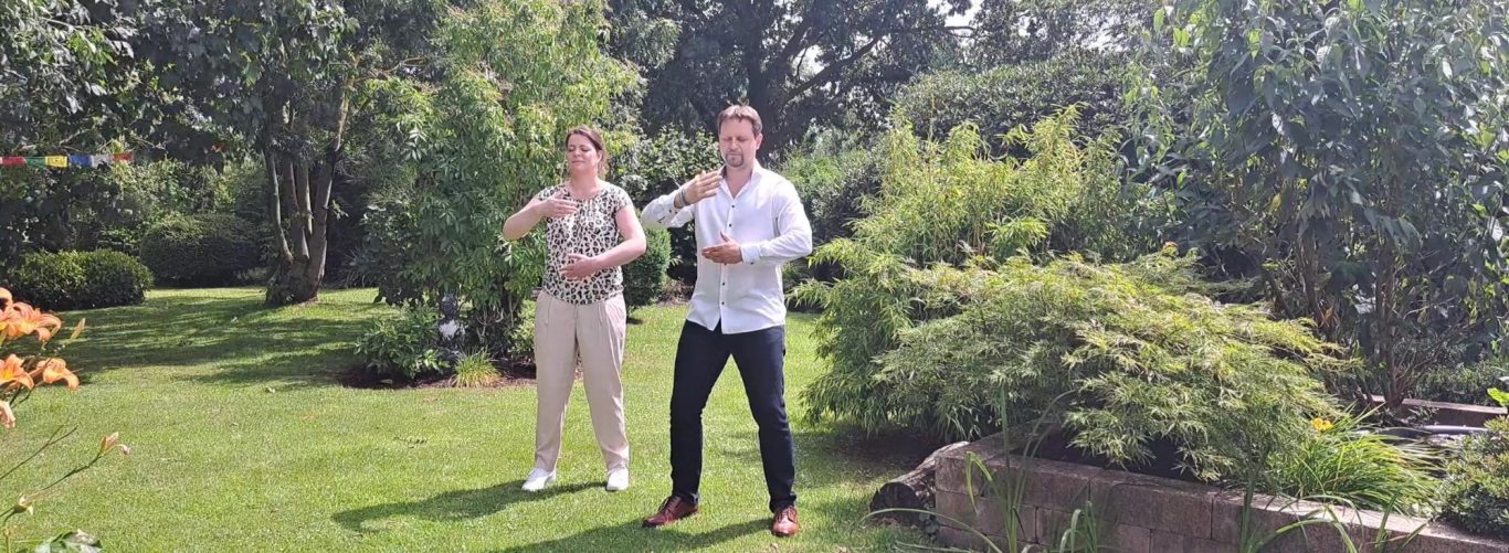 Zwei Personen stehen in einem grünen Garten und posieren fröhlich.