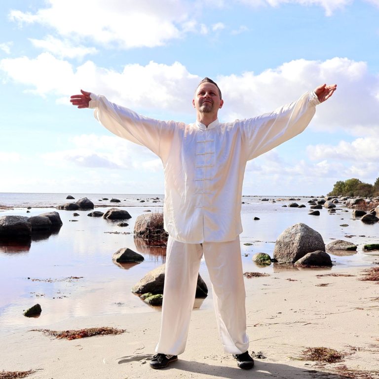 Mann in weißer Kleidung steht am Strand mit ausgebreiteten Armen, umgeben von Steinen.